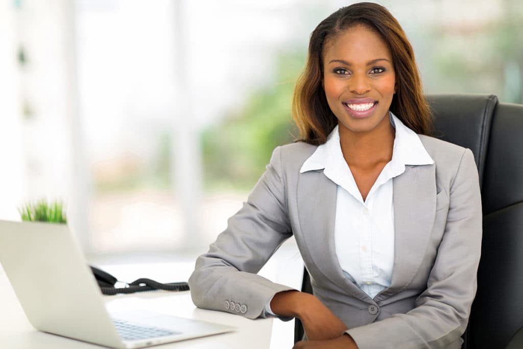 Smiling finance professional in a grey suit seated at a desk with a laptop
